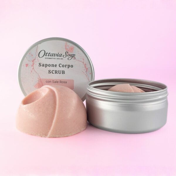 In primo piano sapone tondo colore rosa di fianco a scatolina alluminio aperta. Sullo sfondo il coperchio della scatola con l'etichetta identificativa