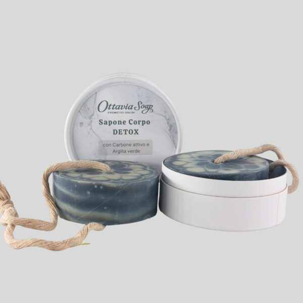 In primo piano un sapone tondo color nero e grigio chiaro. Di fianco, una scatolina tonda in cartoncino bianco. In secondo piano il coperchio con l'etichetta
