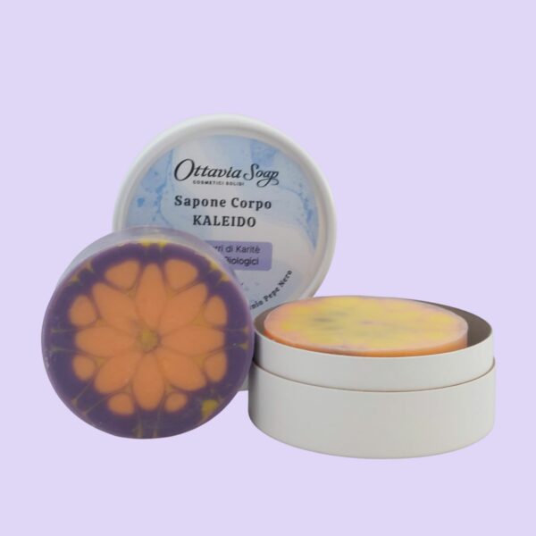 In primo piano un sapone tondo color viola, giallo ed arancione. Di fianco, una scatolina tonda in cartoncino bianco. In secondo piano il coperchio con l'etichetta