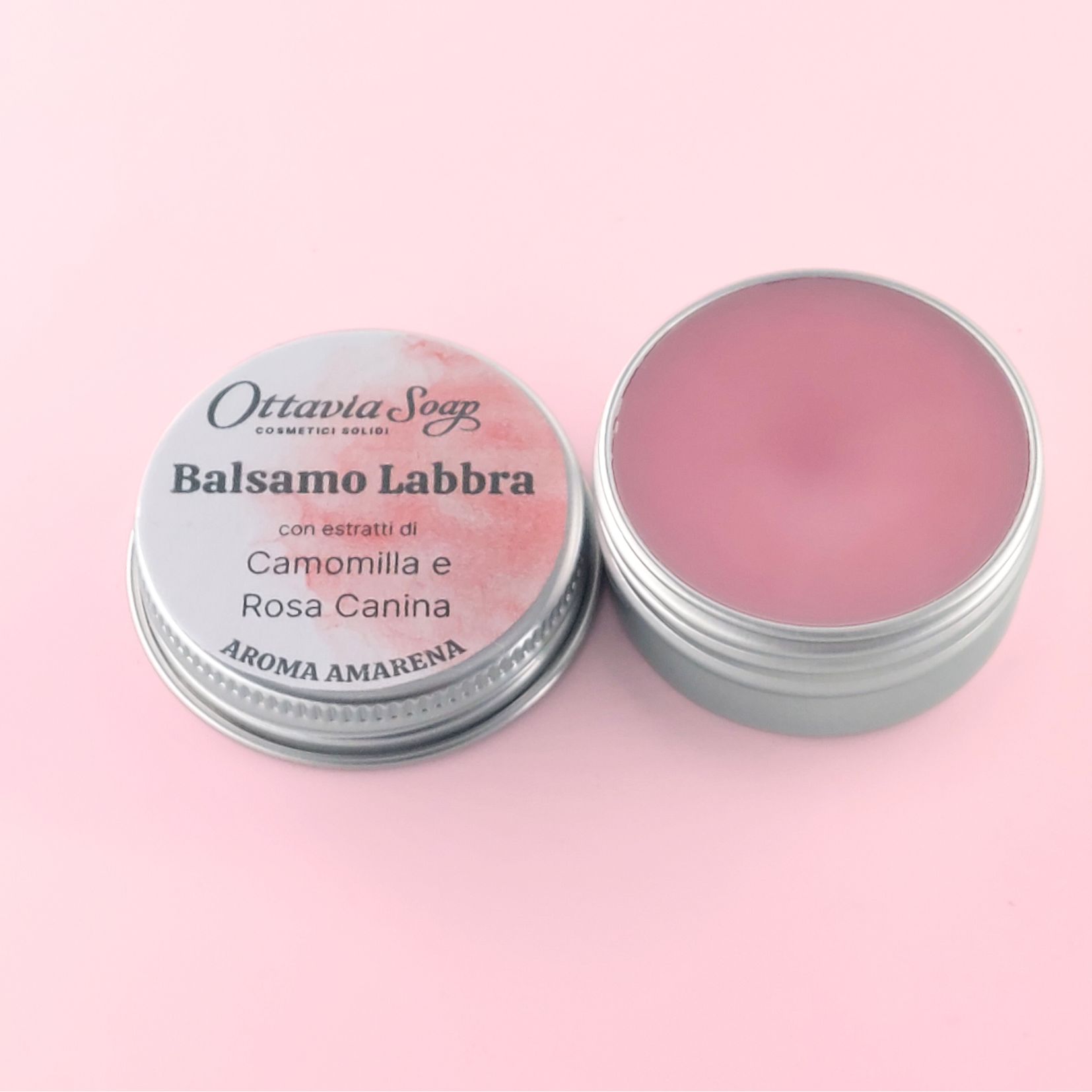 BALSAMO LABBRA Aroma Amarena – (Estratti di Camomilla e Rosa Canina) In primo piano un vasetto in alluminio aperto. Contiene il Burro per le Labbra. Di fianco, il tappo del vasetto con il nome del prodotto e l’etichetta color Rosa