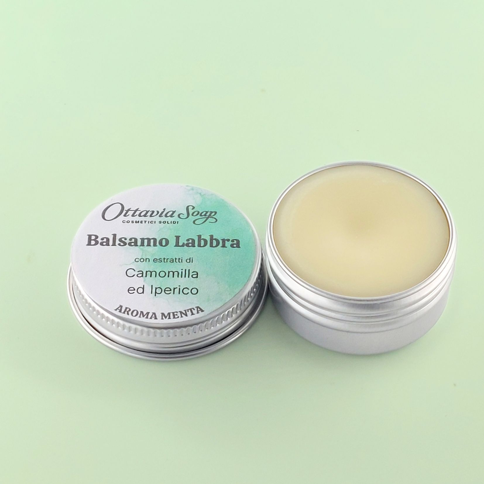 BALSAMO LABBRA Aroma Menta – (Estratti di Camomilla ed Iperico) In primo piano un vasetto in alluminio aperto. Contiene il Burro per le Labbra. Di fianco, il tappo del vasetto con il nome del prodotto e l’etichetta color Verde