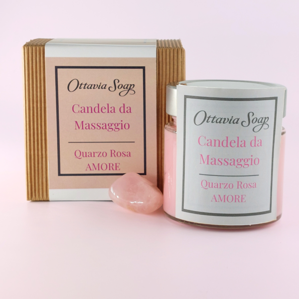 In primo piano un vasetto di vetro della candela da massaggio color rosa. Di fianco al vasetto una pietra di Quarzo Rosa. Sullo sfondo un cofanetto color Avana chiuso con una fascia contenente le informazioni ed il nome del prodotto.