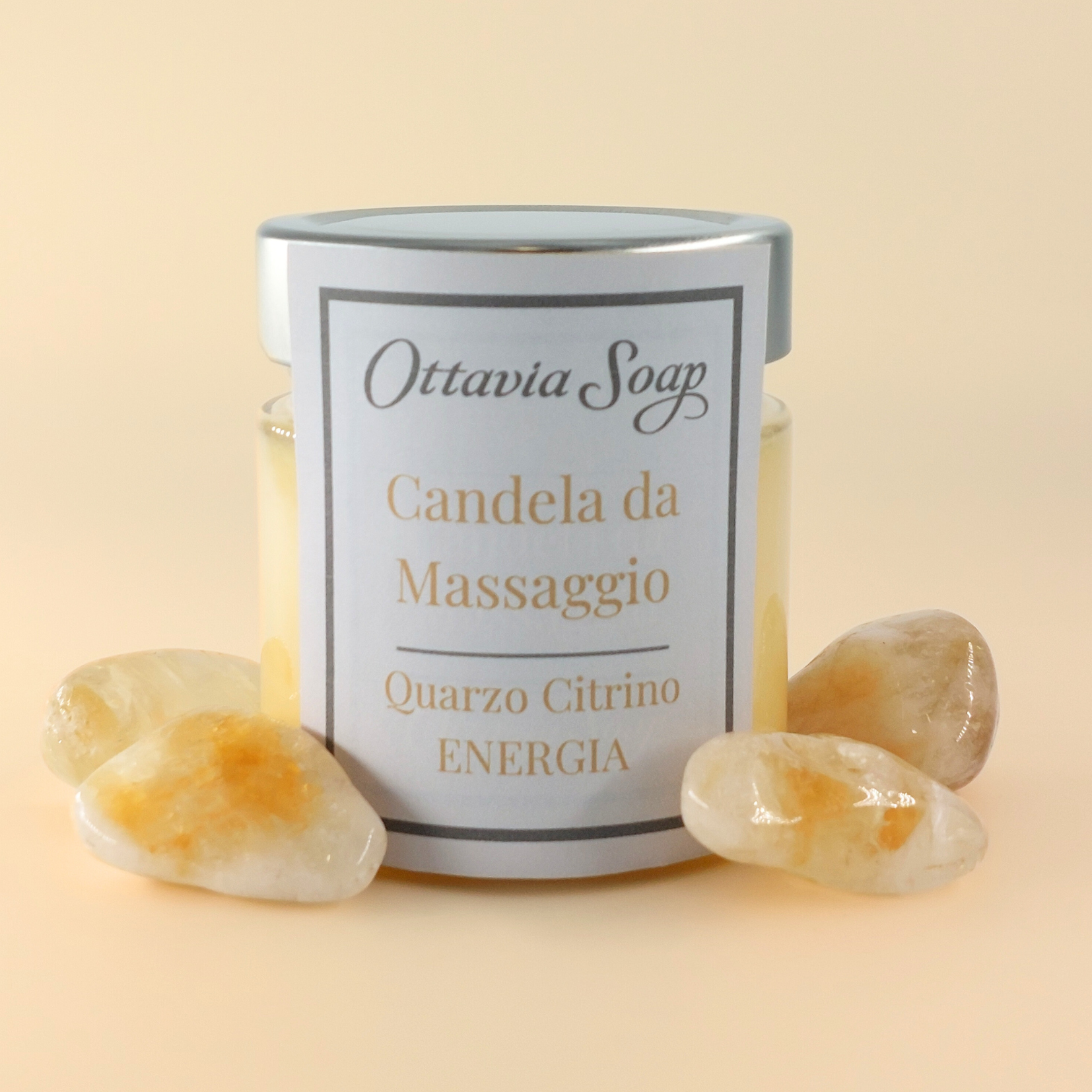 Candela Olio Macadamia e Pietra Quarzo Citrino In primo piano un vasetto di vetro della candela da massaggio color giallo. Di fianco al vasetto una pietra di Quarzo Citrino. Sullo sfondo un cofanetto colora Avana chiuso con una fascia contenente le informazioni ed il nome del prodotto.