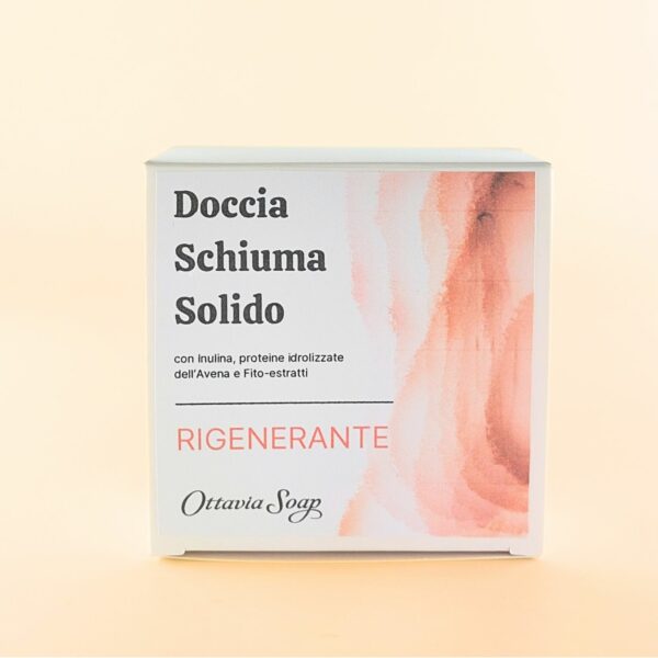Scatolina quadrata in cartoncino bianco posta in verticale. Sul lato frontale una immagine composta da grafica rosa salmone sfumata sul lato destro. Dalla parte sinistra il seguente testo: Doccia Schiuma Solido Rigenerante con Inulina