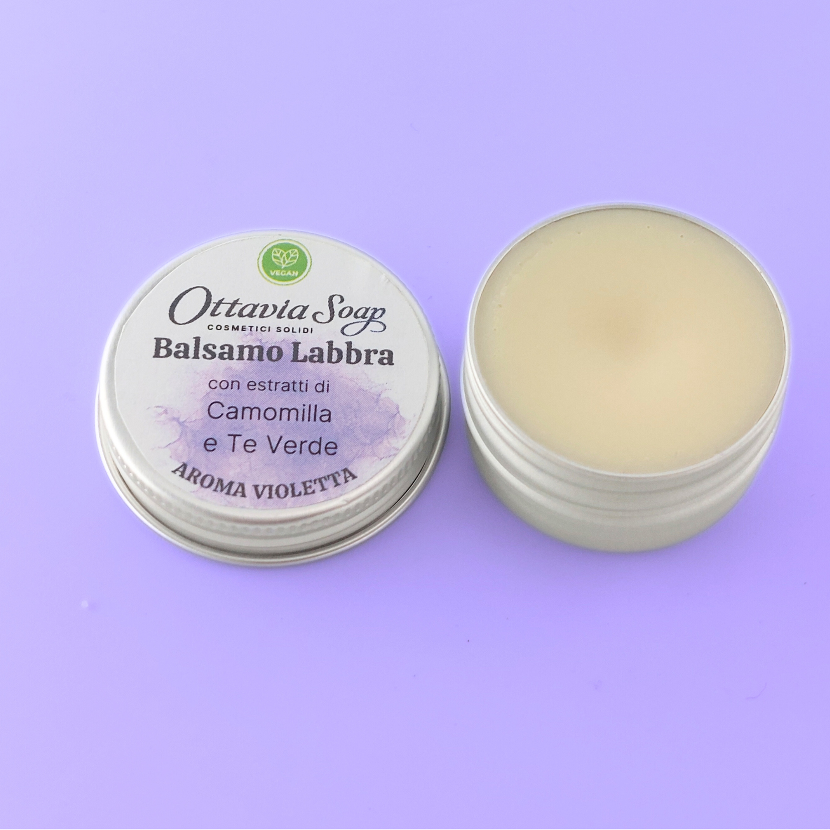 Balsamo Labbra Veg Aroma Violetta Piccolo vasetto tondo in alluminio. E' aperto, sulla sinistra il tappo con l'etichetta, a destra il vasetto di burro solido di colore bianco
